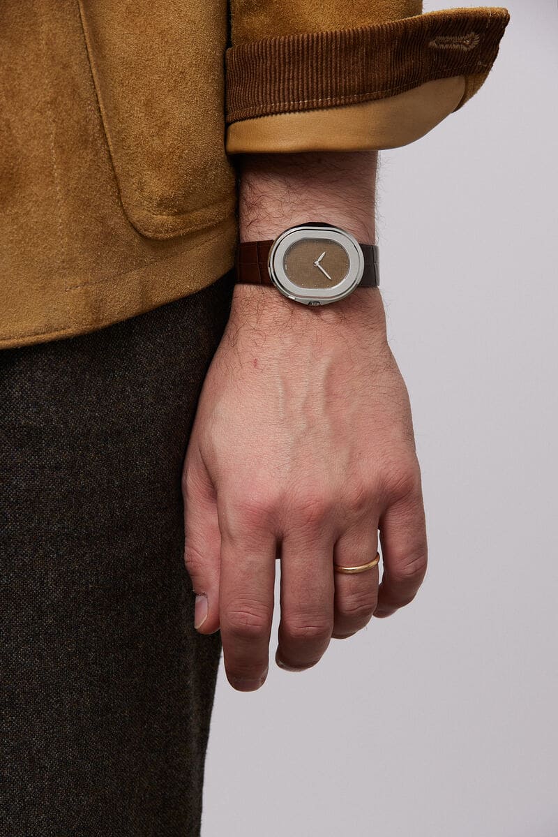 Men’s luxury Serica Parade 1174 stainless steel oval dress watch with brown linen textured dial and brown leather strap on wrist, minimalist Swiss timepiece styled with suede jacket and tailored trousers